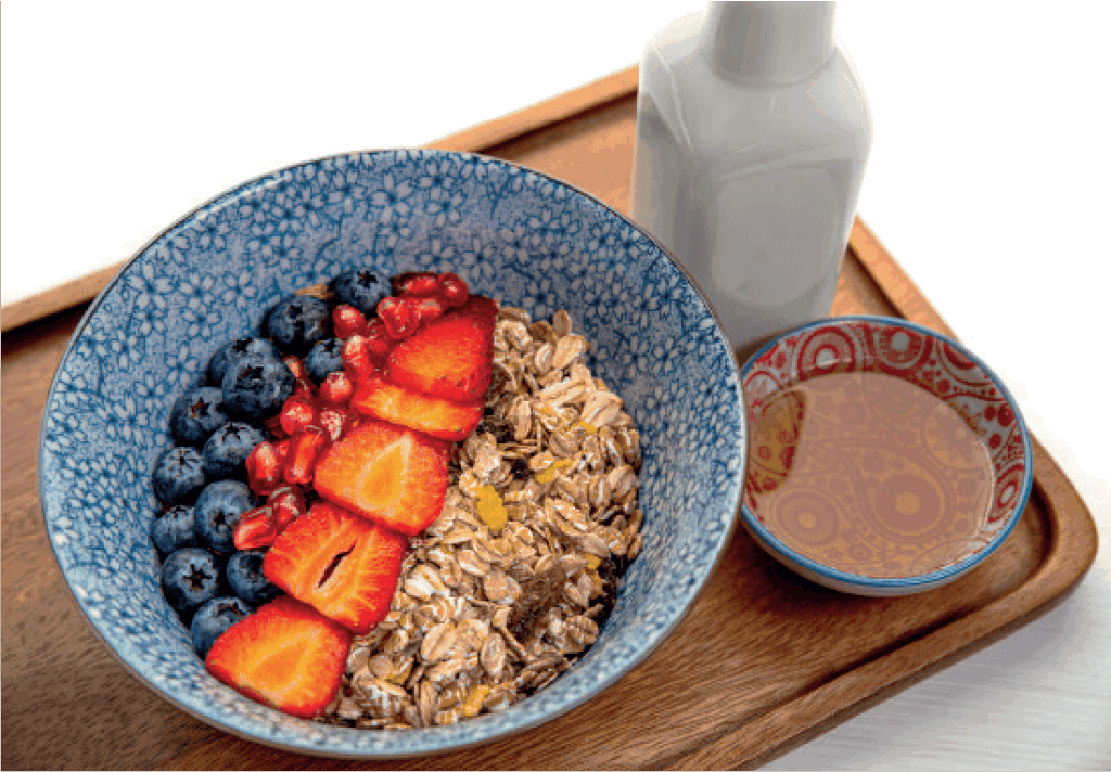 A bowl of muesli with fresh strawberries, blueberries, and pomegranate seeds is served with a side of honey and a bottle of milk.