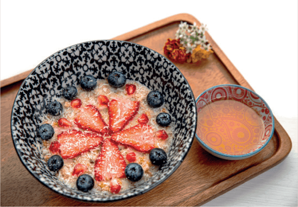 A bowl of creamy oatmeal is artfully topped with sliced strawberries, blueberries, pomegranate seeds, and coconut flakes, with a side of honey.