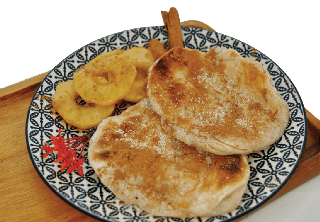Two fluffy pancakes topped with cinnamon sugar are served with cooked apple slices and a red flower garnish on a patterned plate.