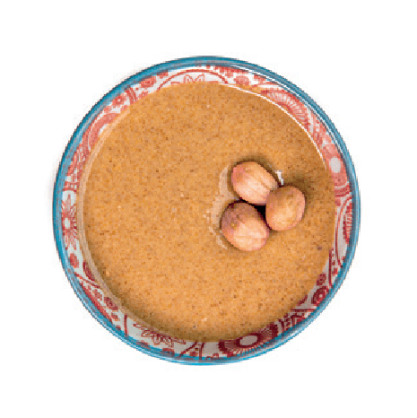 A smooth, light brown peanut butter is topped with three whole hazelnuts in a small patterned bowl.