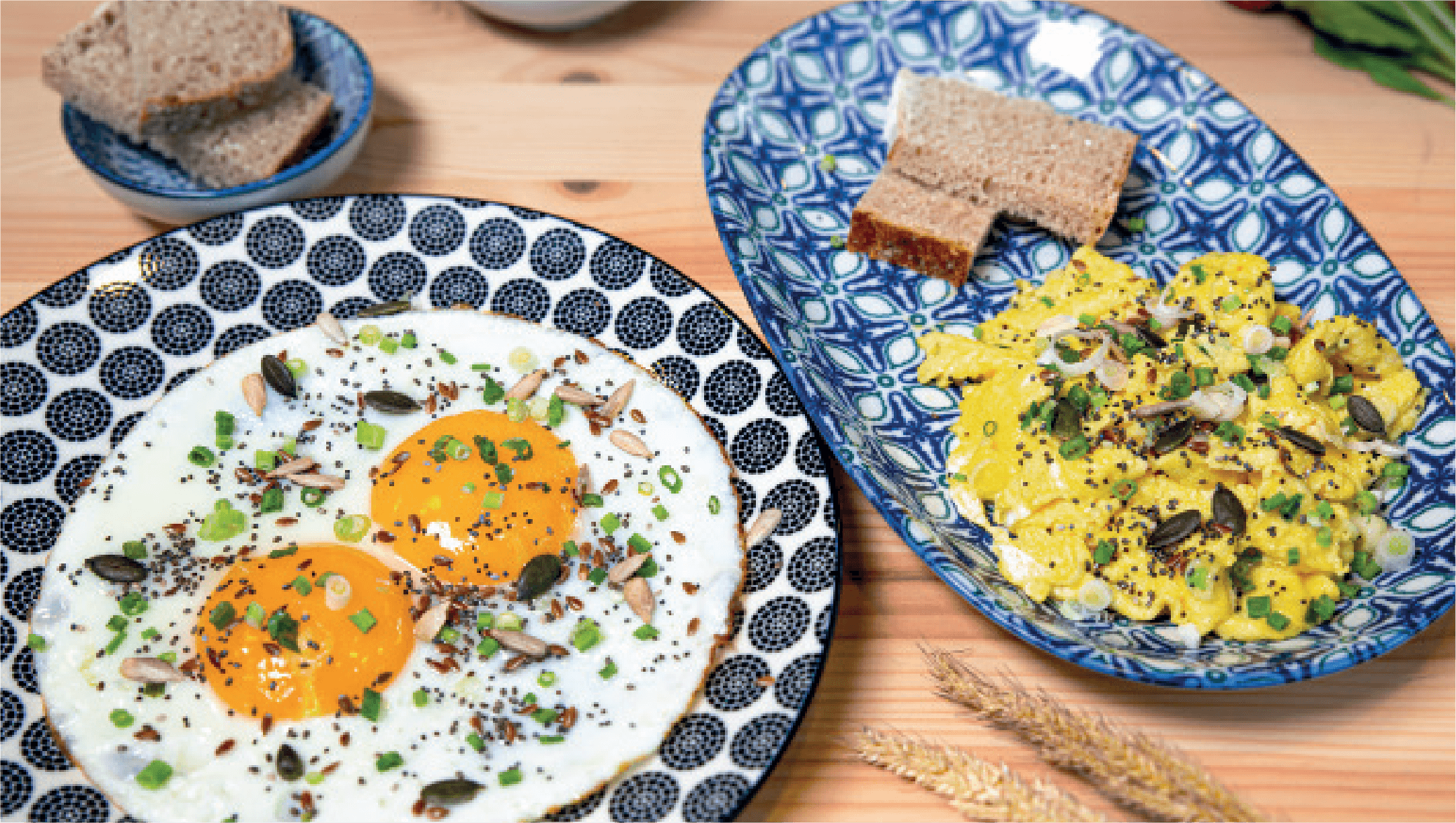 Two sunny-side-up fried eggs topped with seeds and chives, and a separate dish of scrambled eggs with similar toppings, are served with rye bread.