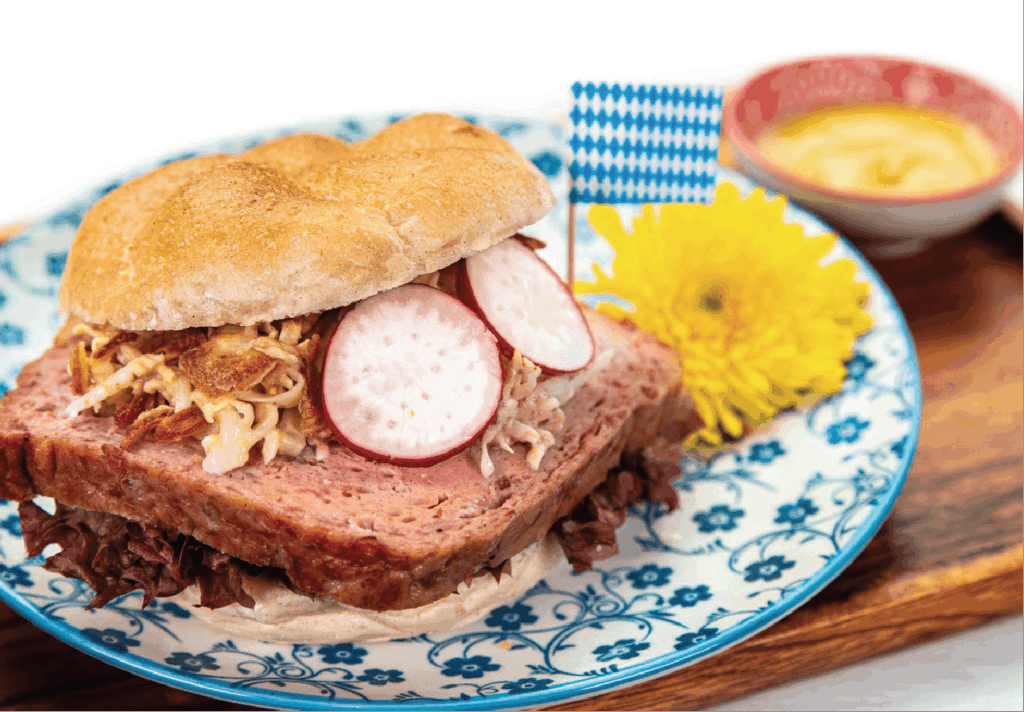 A German meatloaf (Leberkäse) sandwich with sauerkraut and radish slices is served on a patterned plate with a Bavarian flag pick.