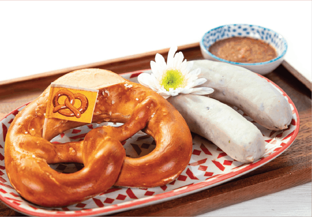 Two Weisswurst sausages, a large pretzel with a pretzel flag, and a side of sweet mustard are presented on a patterned plate.