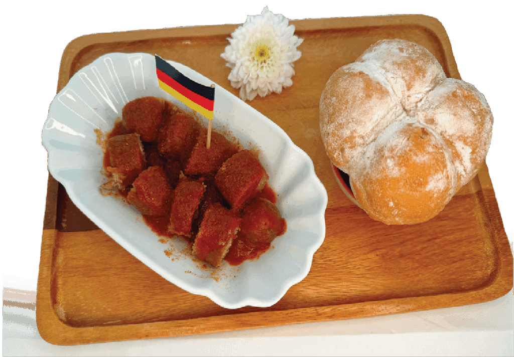 Sliced currywurst sausage topped with curry ketchup and powder is served in a paper dish with a German flag pick and a bread roll on a wooden tray.