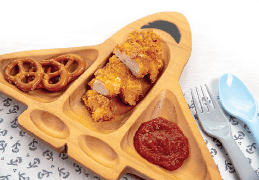 Chicken nuggets, pretzels, and ketchup are arranged on an airplane-shaped wooden plate for a child's meal.