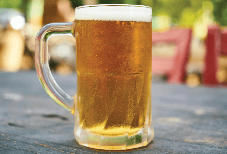 A frosty glass mug filled with amber beer and a foamy head sits on an outdoor wooden table.
