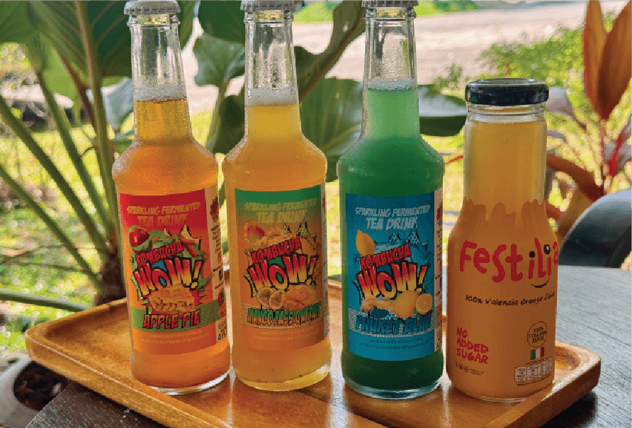 Three bottles of Kombucha WOW! in various flavors and one bottle of Festilia orange juice are arranged on a wooden tray.