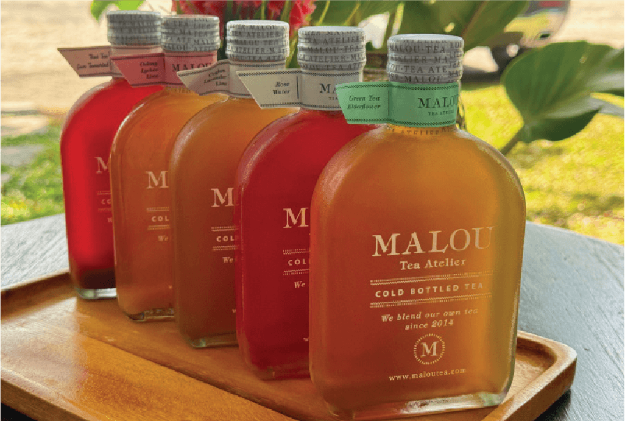 A lineup of colorful MALOU Tea Atelier cold bottled teas is displayed on a wooden tray.