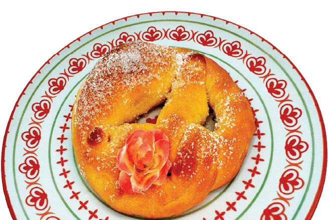 A sweet pretzel dusted with powdered sugar and adorned with a pink edible flower sits on a decorative plate.