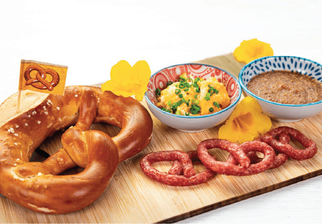 A soft pretzel is accompanied by two dipping sauces and pretzel-shaped cured meats on a wooden board.