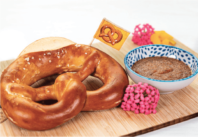 A classic soft pretzel with coarse salt is served on a wooden board alongside a bowl of brown dip and pink flowers.
