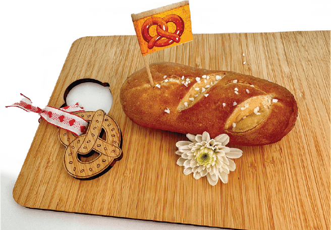 A salted pretzel stick garnished with a small pretzel flag and a white flower rests on a wooden cutting board.