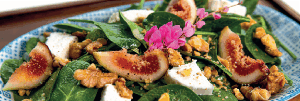 A fresh spinach salad features sliced figs, chunks of white cheese (likely feta), walnuts, and a pink flower garnish.