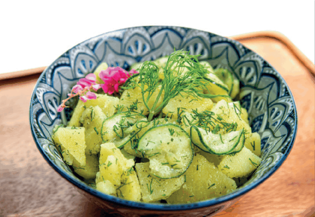 A German-style potato salad with sliced cucumbers and fresh dill is served in a patterned blue and white bowl.