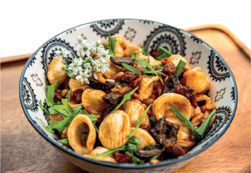 A bowl of orecchiette pasta is tossed with greens, sun-dried tomatoes, pine nuts, and possibly mushrooms, garnished with white flowers.