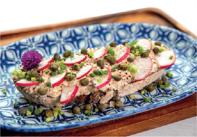An open-faced sylt sandwich on dark bread features a creamy tuna spread topped with radish slices, capers, and chives.