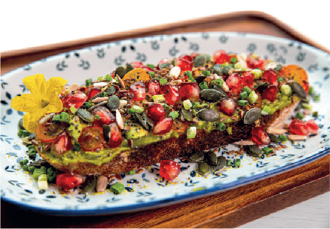 Avocado toast generously topped with pomegranate seeds, pumpkin seeds, and chives is served on a floral plate.