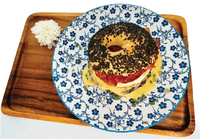 A black sesame seed bagel sandwich filled with bacon, egg, and cheese, drizzled with sauce, is presented on a floral plate on a wooden tray.