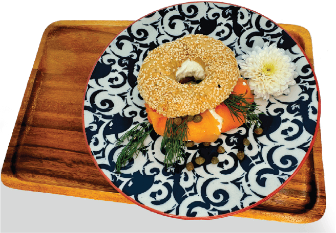 An open-faced sesame seed bagel with lox, cream cheese, dill, and capers is served on a patterned plate atop a wooden tray.