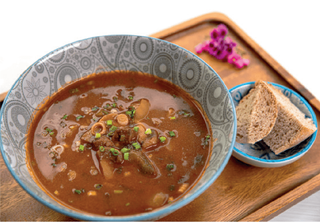 A dark, savory goulash soup with chopped green onions is served in a patterned bowl with slices of bread on a wooden tray.