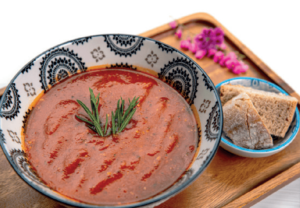 A rich red tomato soup, garnished with rosemary, is served in a patterned bowl with slices of bread on a wooden tray.