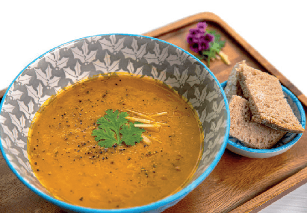 A bowl of orange carrot soup, garnished with cilantro and ginger, is served with slices of brown bread on a wooden tray.