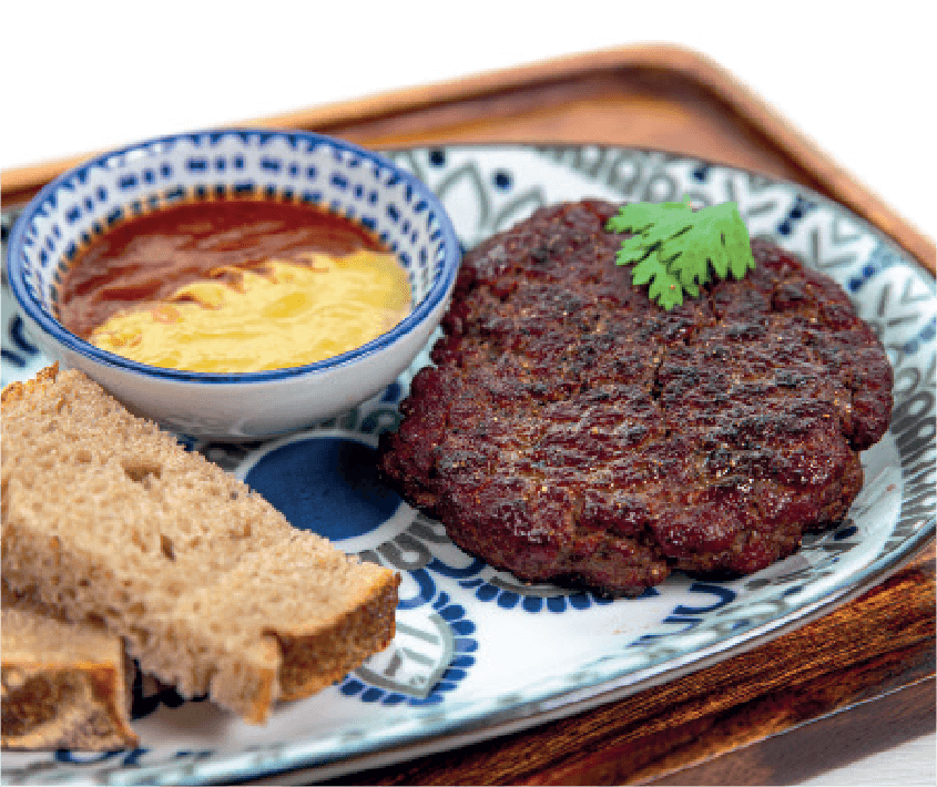 A grilled beef patty is served with two slices of rye bread and a side of dipping sauce (possibly mustard and ketchup) on a patterned plate.