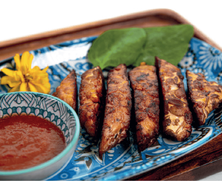 Several strips of grilled tempeh are arranged on a blue patterned plate with a side of red dipping sauce and a yellow flower garnish.