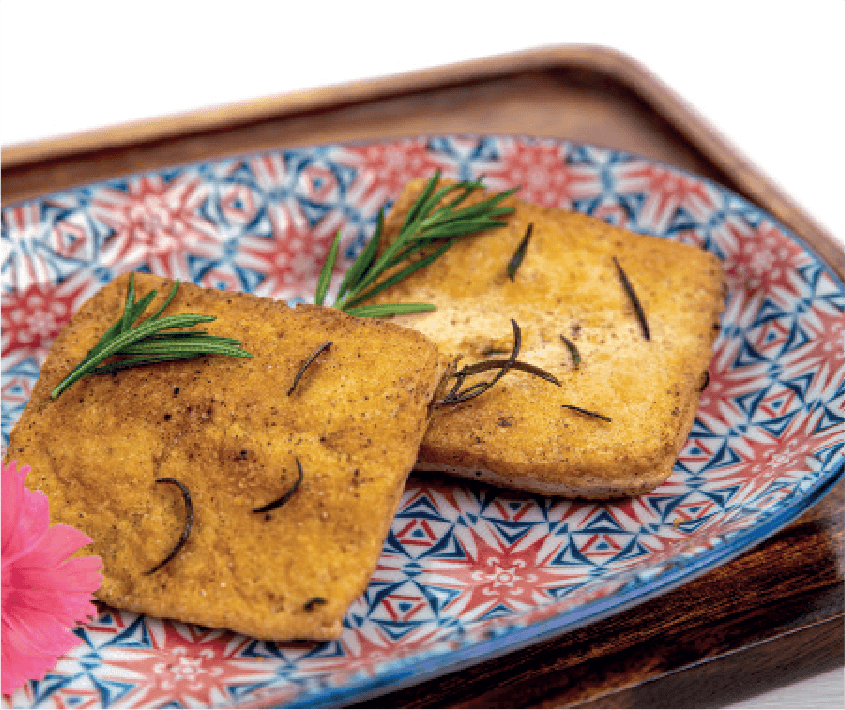 Two square, golden-brown pieces of seasoned tofu are garnished with rosemary sprigs on a colorful patterned plate.