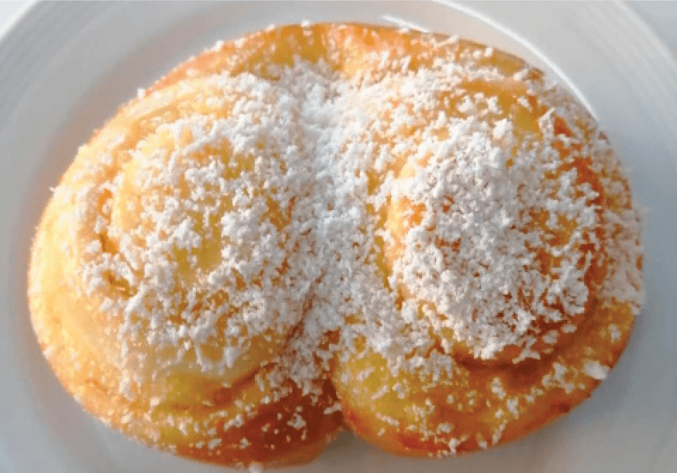 A golden, swirled pastry, possibly a Franzbrötchen or similar, is dusted with powdered sugar on a white plate.