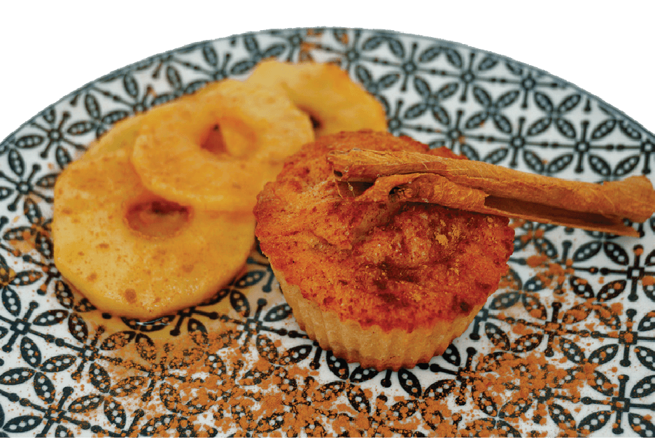 An apple muffin with baked apple slices and a cinnamon stick garnish is presented on a patterned plate dusted with cinnamon.