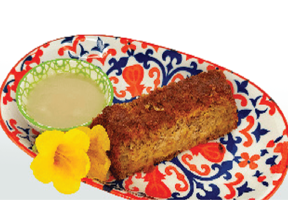 A slice of moist-looking bread, possibly banana bread, is served with a side of white dipping sauce and a yellow flower garnish on a colorful patterned plate.