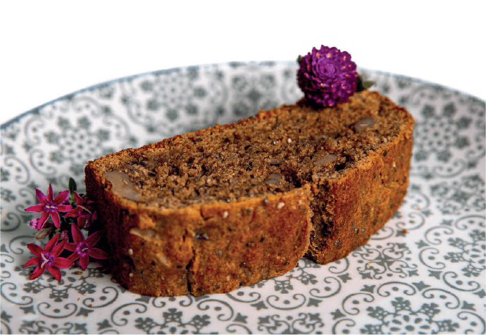 A slice of dark, dense nut and seed bread is presented on a patterned plate with a small flower garnish.